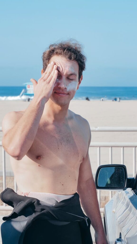 Shirtless man applying sunscreen at a sunny beach, emphasizing skin protection.